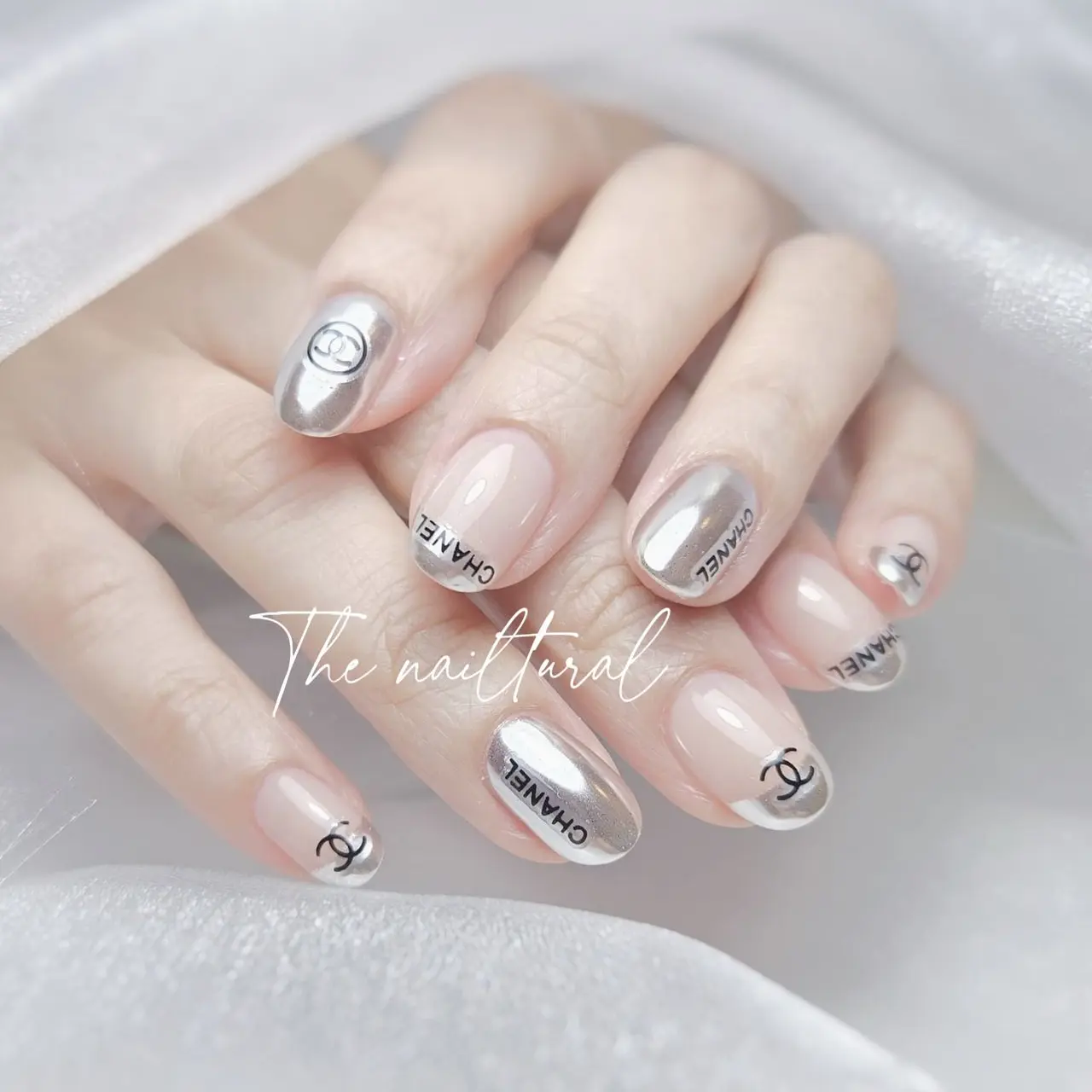  oval-shaped nails ,  chic and branded nail art design,  nude base ,CHANEL reflective silver chrome ,  "CC" Chanel logo i fashionable ,  luxurious nail design
