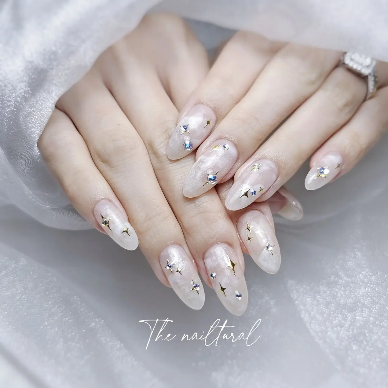 almond-shaped nails,  celestial-inspired design, a sheer, milky white base, tiny, sparkling rhinestones shapes,  stars, small round gems. Delicate gold star-shaped decals.   ethereal and glamorous nail art.